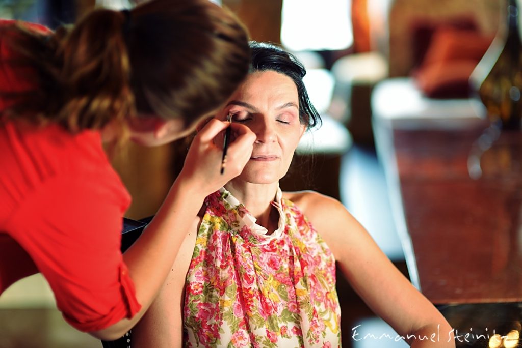 Scène en coulisses illustrant la préparation d’un shooting photo portrait professionnel, focus sur la section maquillage et coiffure. Une maquilleuse applique le maquillage des yeux sur une femme assise, dans une ambiance chaleureuse et intimiste, avec une profondeur de champ réduite et un arrière-plan flou. Image idéale pour illustrer un article sur la préparation avant séance photo, le styling, la mise en beauté, l’organisation d’un shooting et les services de beauté professionnels.
