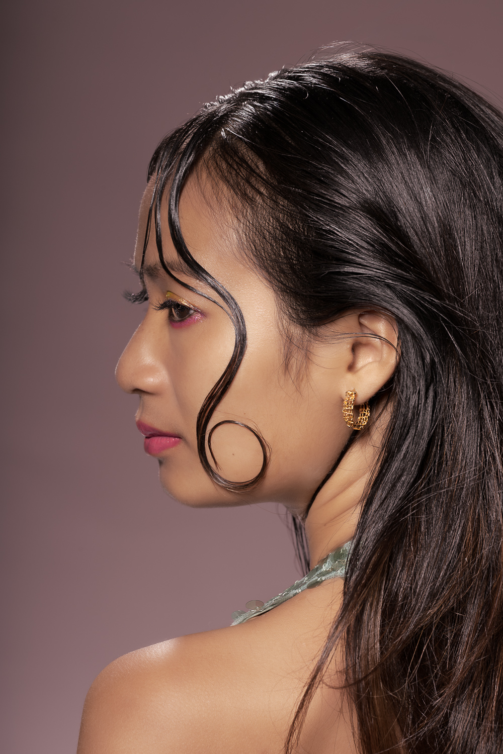 Portrait studio raffiné d’une jeune femme vue de profil avec cheveux brillants, mèche bouclée, maquillage subtil et boucle d’oreille dorée sur fond rose poudré. Idéal pour illustrer la beauté, la mode, les cosmétiques, le soin de la peau et l’élégance féminine.