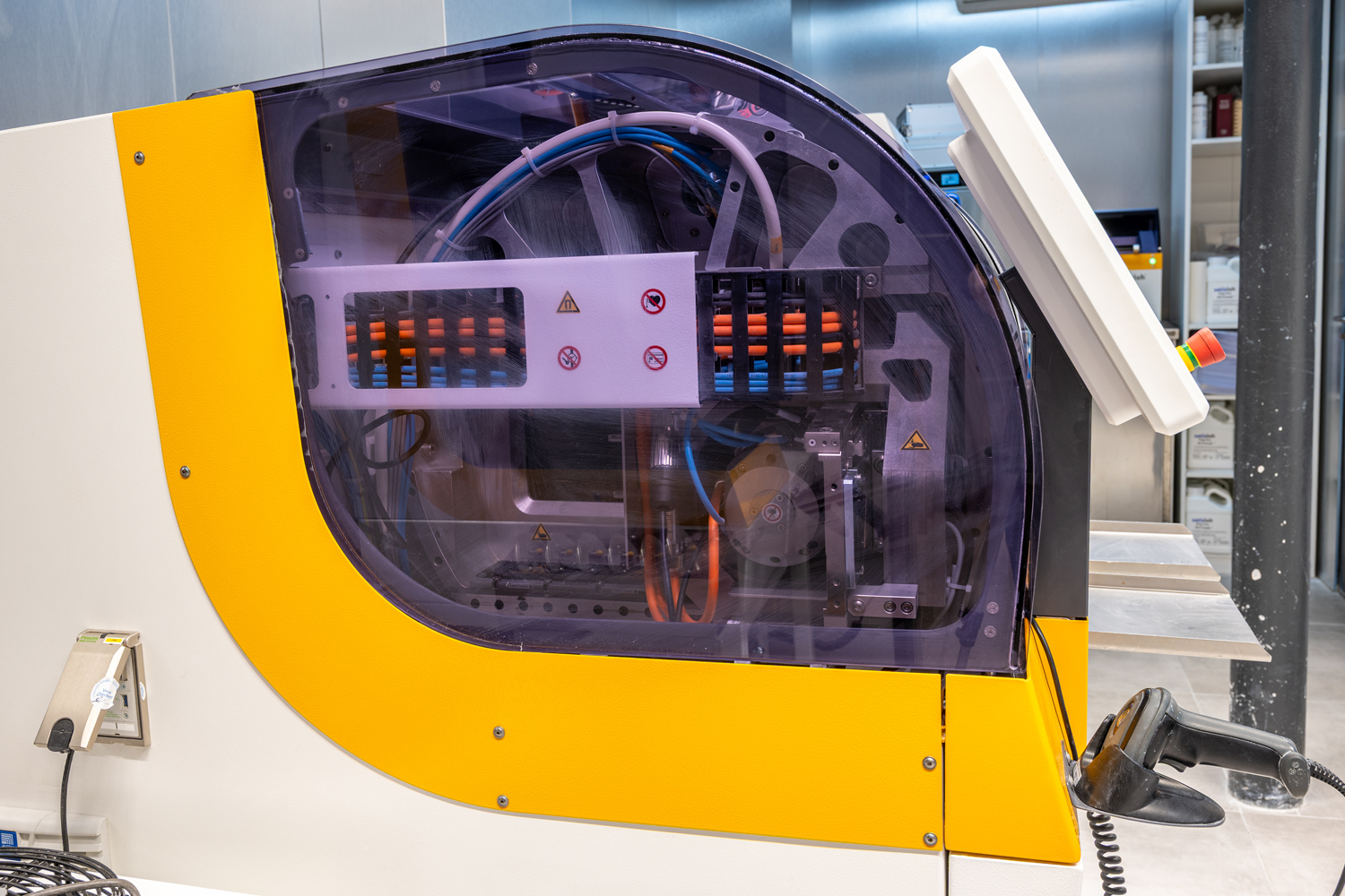 Photo horizontale, cadrage serré sur une machine industrielle, environnement de laboratoire moderne, absence de personnes, capot transparent violet, structure jaune et blanche, câbles internes visibles, lecteur manuel au premier plan, lumière artificielle, ambiance propre, technique et professionnelle.